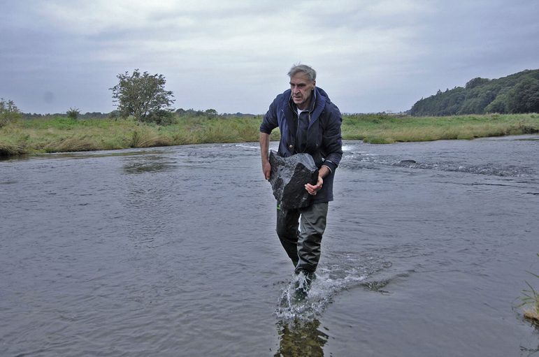 A man with short grey hair wearing dark clothes carrying a large grey rock as he walks through a shallow body of water