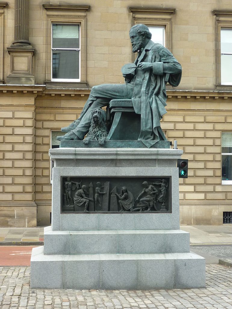 A greenish grey statue of a man with facial hair and a long jacket sitting on a chair. The statue sits on top of a tired stone plinth.