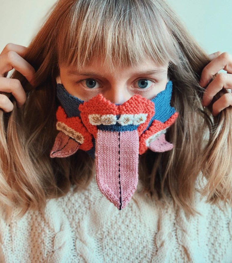 A woman with long hair wearing a cream knitted jumper and a knitted facemask featuring three red mouths with tongues sticking out.