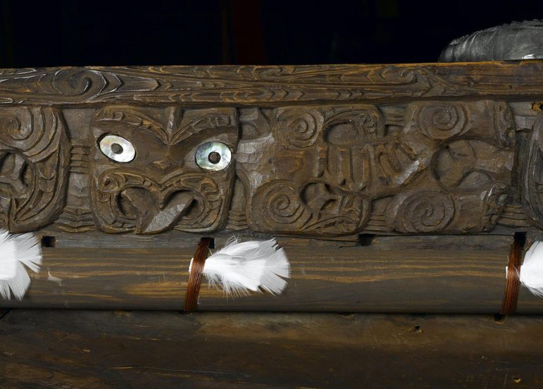 A detail of a wooden canoe with carved faces and feathers attached with brown straps
