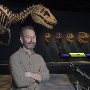 A bearded man standing smiling in front of a dinosaur skeleton