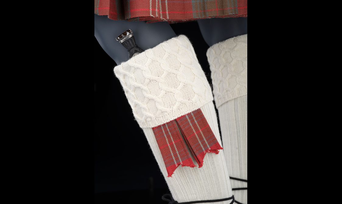 A close up of a mannequin wearing white wool socks with tartan tassels and a small dagger tucked into the sock cuff.