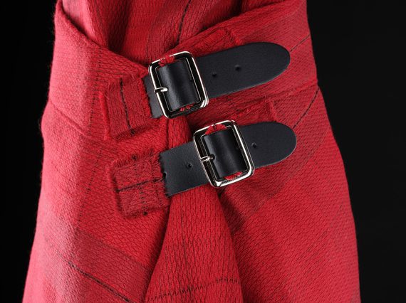 A detail of a red kilt with a subtle grid pattern with two leather buckles fastening the side.