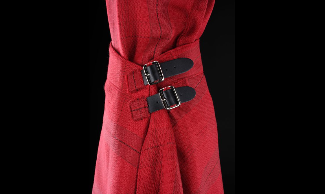 A detail of a red kilt with a subtle grid pattern with two leather buckles fastening the side.