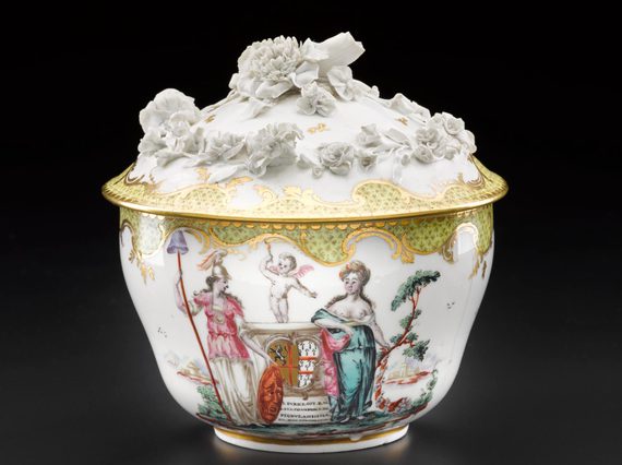 An elaborate sugar bowl with a lid. It is made of porcelain and it is decorated with people, a cherub, and a carriage and flowers in relief on the lid.