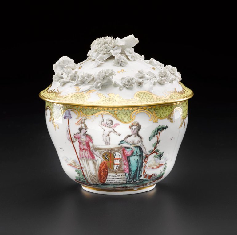 An elaborate sugar bowl with a lid. It is made of porcelain and it is decorated with people, a cherub, and a carriage and flowers in relief on the lid.