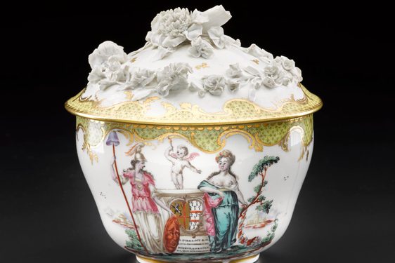 An elaborate sugar bowl with a lid. It is made of porcelain and it is decorated with people, a cherub, and a carriage and flowers in relief on the lid.