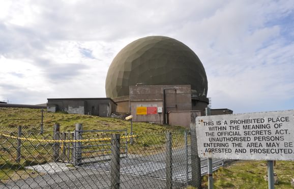 A large round building that looks a bit like a gold ball, behind a chain link fence with a warning sign outside that prohibits unauthorised entrants.