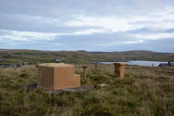 Brick block buildings sat in a field in Shetland, with the sea in the background and cloudy skies above.