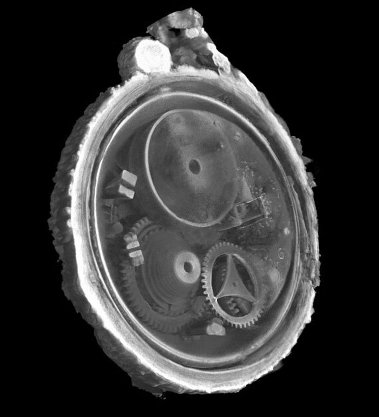 A black and white scan showing the insides of a circular watch at an angle, including a cog on the middle left, a circular plate with a hole ion it at the top left, and a round rim with a curved triangular support in the middle. The outside of the watch shows a bumpy outer crust