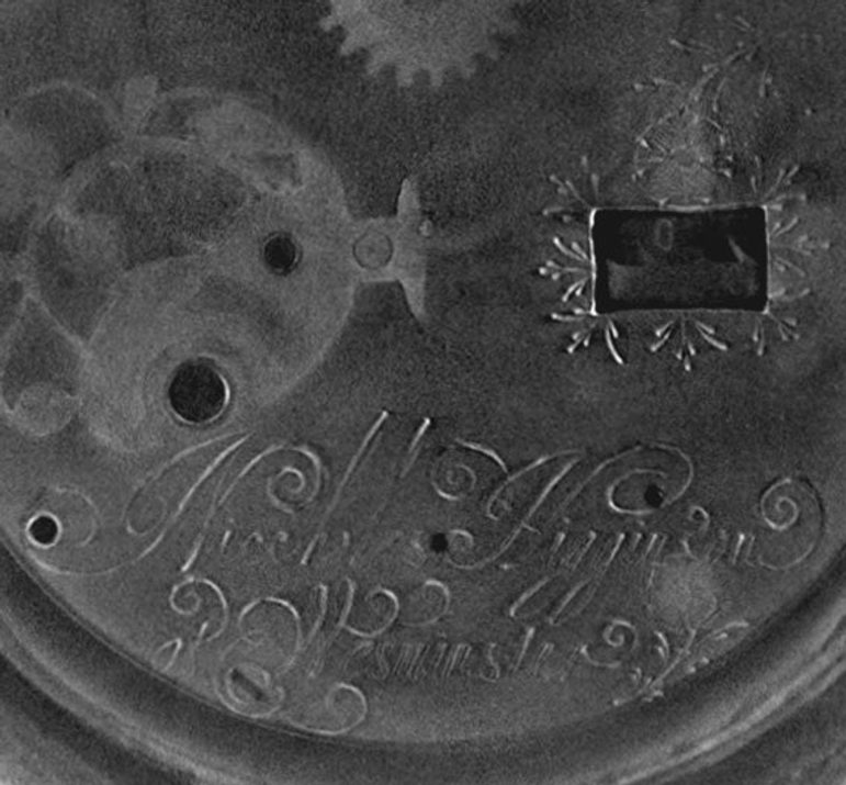 A black and white scan showing the insides of a circular watch close up and from the back. There is an engraving on the bottom middle. There is also a ghostly image of the cogs and wheels that are in the middle of the watch. The outside of the watch shows a bumpy outer crust