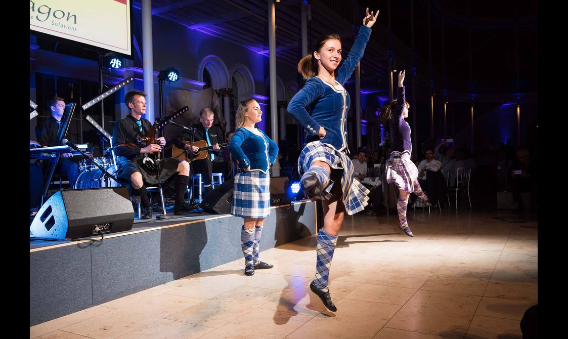 Scottish higland dancers in blue outfits dancing infront of a ceilidh band.