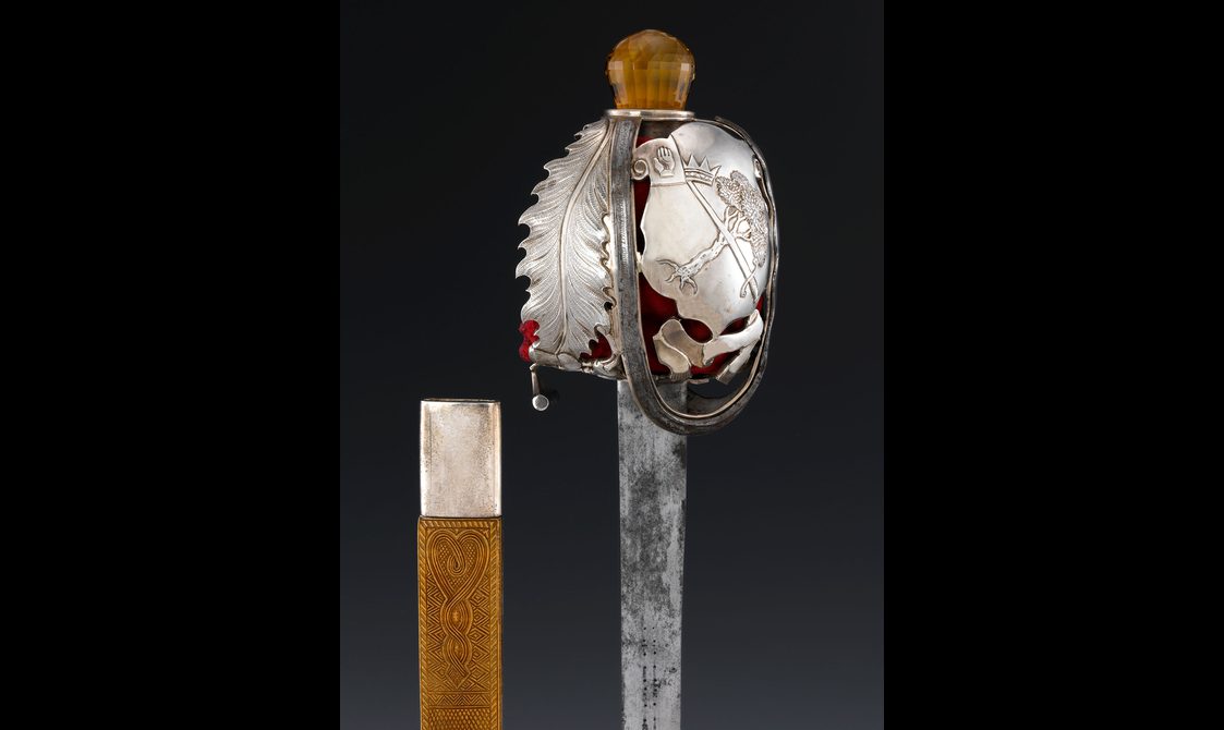 Close up of the hilt of a sword with an engraved leaf and coat of arms.