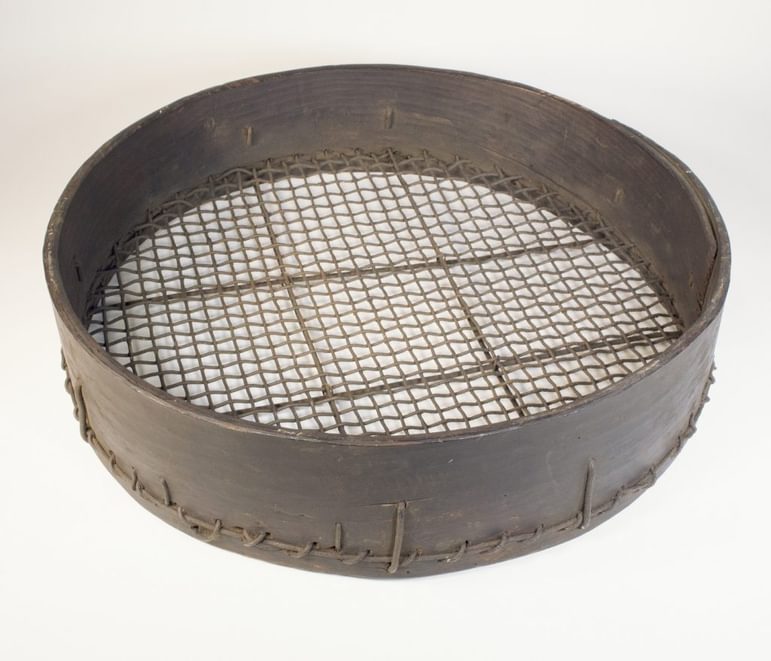 A brown mesh sieve with a thin, high wooden rim laying flat down on a cream-coloured surface.