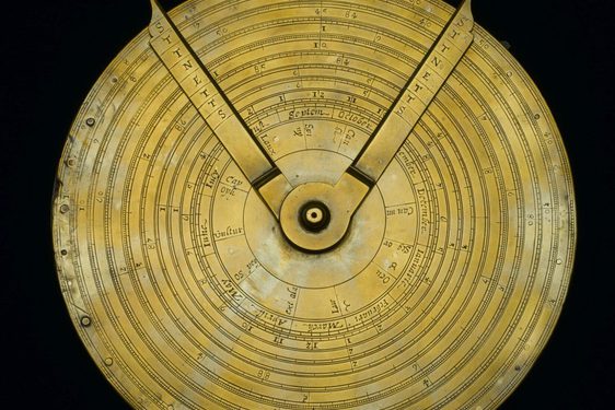 A colour photograph of a yellow metal circular device against a back background. The device has two dials attached at the centre and finishing at the circumference. There are letters engraved on both arms. There are concentric rings on the devoice and engraved measurements.