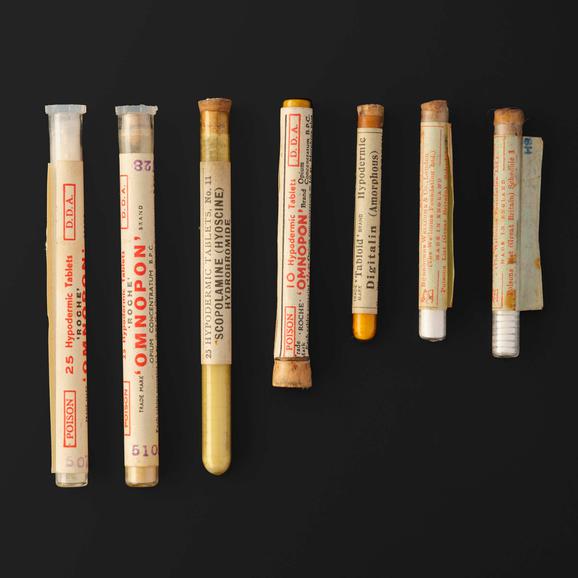Seven glass tubes containing tablets lying in a row. Each tube is wrapped with a label.