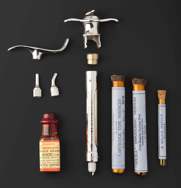 A metal cartridge style syringe next to three glass vials of needles. There is a small red bottle containing anaesthetic solution. There is an extra syringe handle and heads for the needles.
