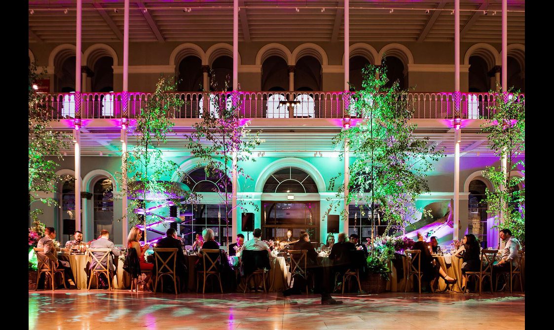 A dinner reception in a large multi-level museum gallery. There are trees lining the dinner table and purple lights illumintaing the space.