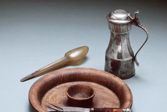 A wooden plate with a knife, fork and spoon as well as a small silver lidded jug.