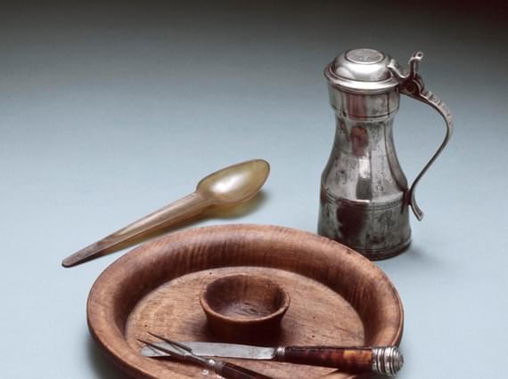 A wooden plate with a knife, fork and spoon as well as a small silver lidded jug.