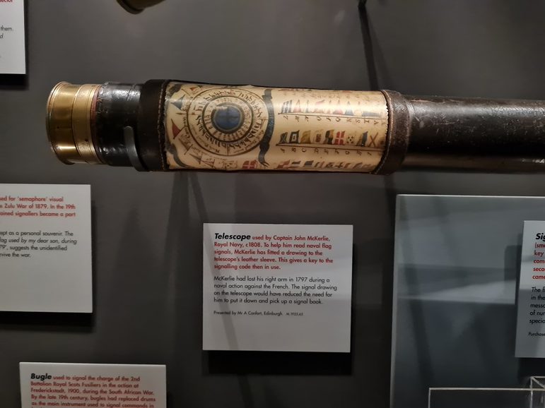 a telescope on display in a museum case, with an explanatory label underneath it. The telescope is black, with gold detailing.