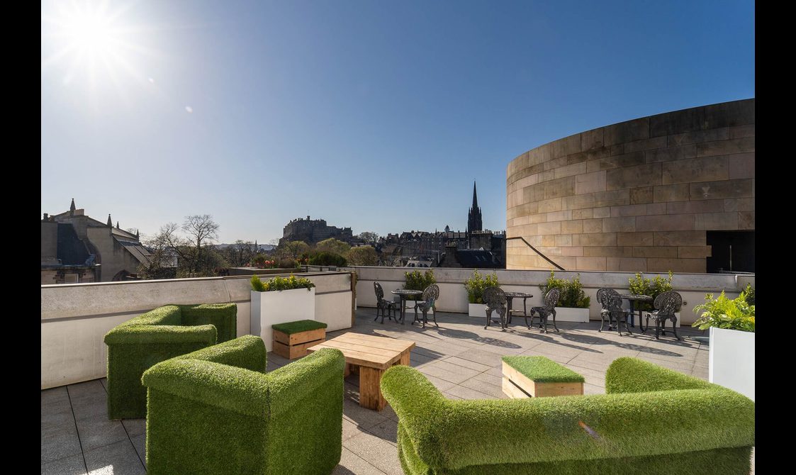 A large, stone terrace with sunny views over to Edinburgh Castle. There are tables, chairs, and large, grass-covered seats spread around.