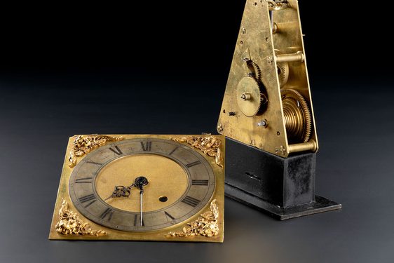 A round clockface on a gold square laying beside a gold triangle with cogs on its surface and its inside. mounted on a rectangle black base.