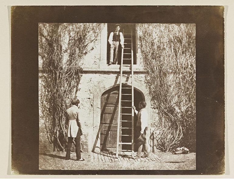 A black and white image of a man in a window of a stone building, with another man on the ground holding a ladder to the window while another man stands on the ground watching. The building has plants growing up its facade.