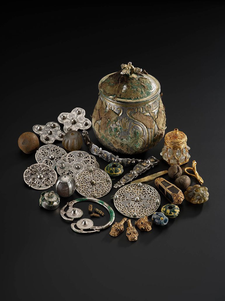 A silver round vessel with a gilded lid engraved with various round and organic shapes surrounded by other items from the Galloway Hoard.