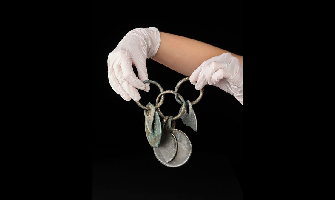 White gloves holding three rings linked together in a row. Five bronze disks are hanging from these rings, creating a rattle. The surface is silvery with some green corrosion.