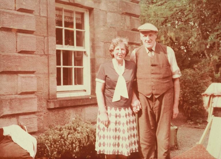A faded photoraph of a man and woman standing holding hands in front of a Georgian stone house.