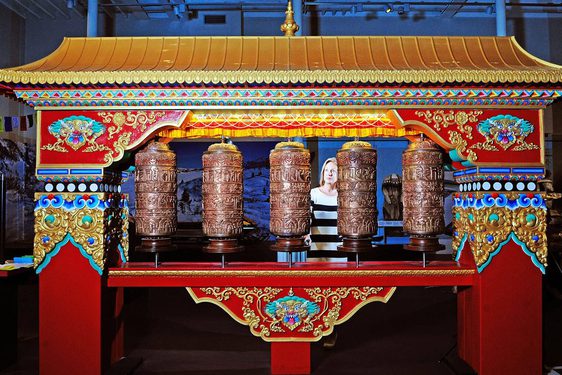 A woman stands behind a large Tibetan structure that is red with a gold roof and decorated pillars. It has five brown spinning prayer wheels at its centre.