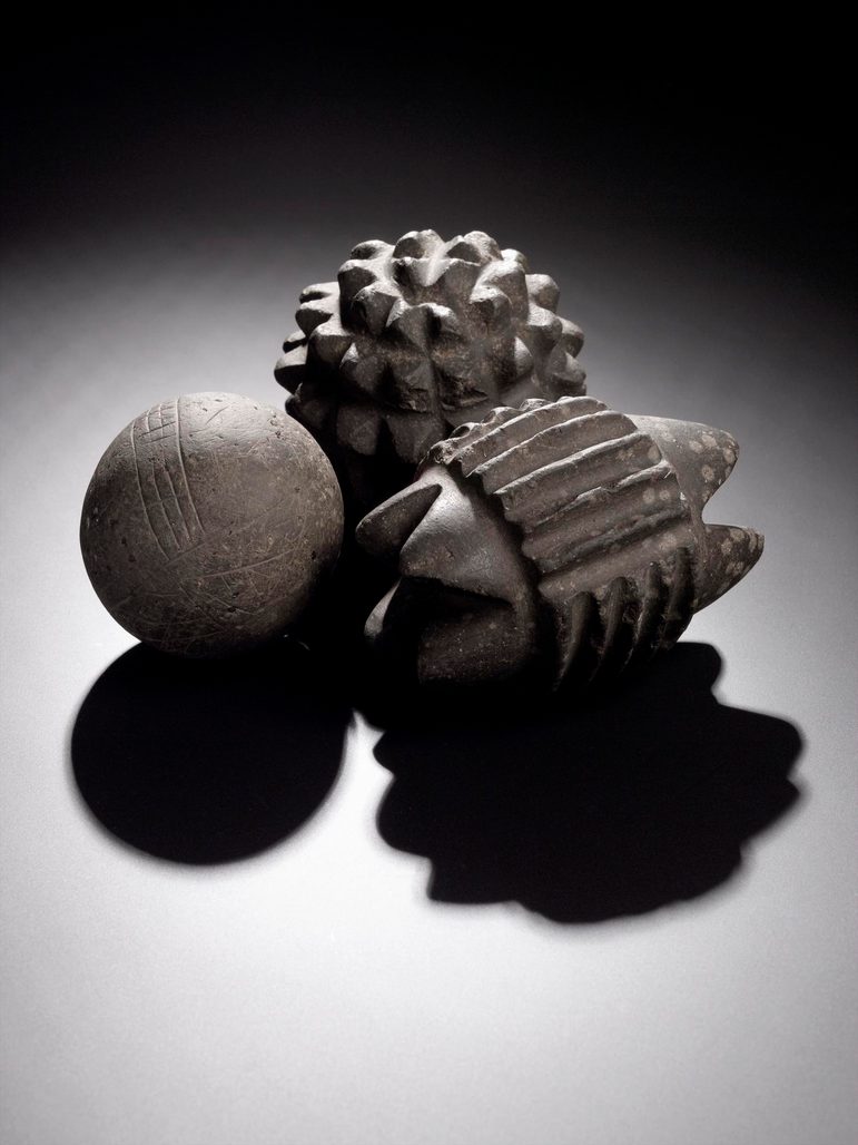 Three grey stone balls, one shaped as a circle with linear decorations, one carved with protruding triangles, and another carved out of triangles to the ends and linear decorations.