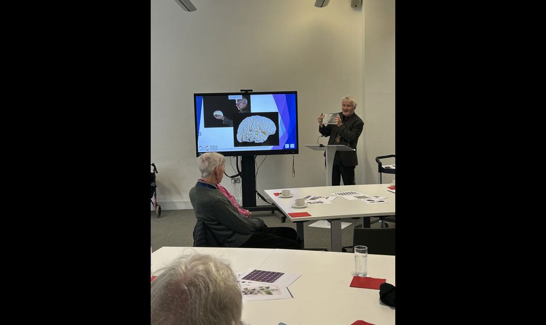 A man stands in front of a group of people giving a presentation. Next to him is an image of a drawing of a brain. 