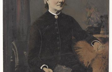 An old photograph of a seated woman in a black Victorian dress buttoned to the top wearing a white and black hat. She is holding a book in one hand and the photograph has been coloured.