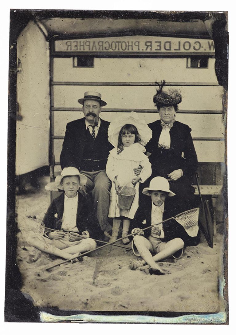 A black and white image of a family with a mother and father and three children at the beach. They are all dressed in Victorian clothes.