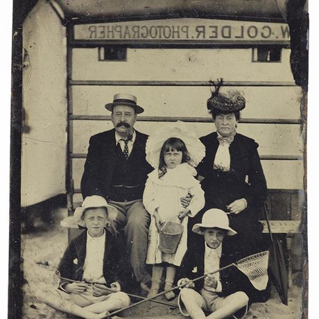 A black and white image of a family with a mother and father and three children at the beach. They are all dressed in Victorian clothes.
