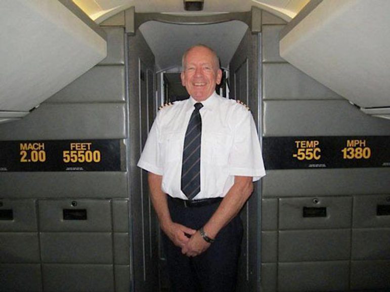 A smiling man in a white short sleeved shirt and tie with pilot uniform markings, standing in a passenger airplane