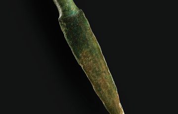 A weathered bronze knife tip with a small hole in one end on a black background