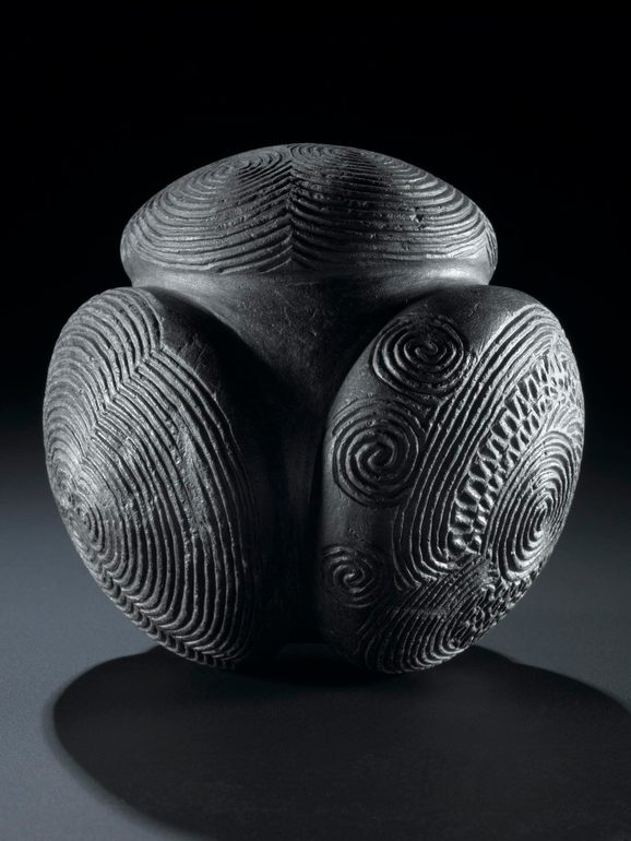 A grey ball with three circular disks carved out of it, decorated in various spiral carvings, set against black background.