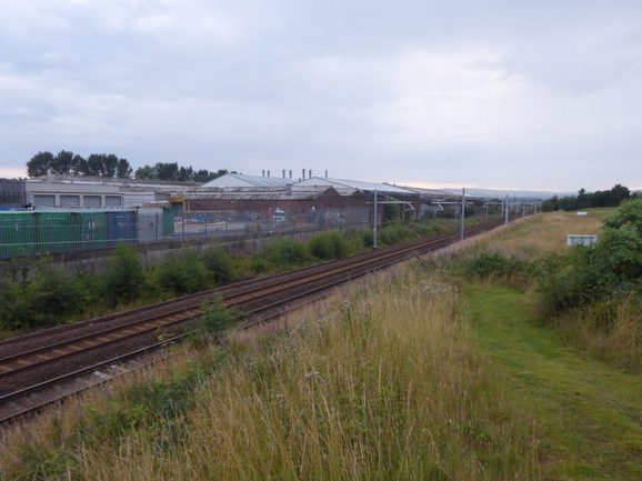 A landscape photograph with grass at the forefront, trains station with train tracks running from left corner to the right.