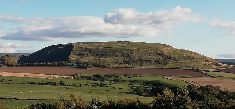 Landscape shop under a blue sky and fading light of a whaleback-shaped hill with layers of old ramparts just barely visible.