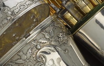 A decorative silver lidded container beside a plain silver container holding various gold objects.