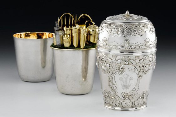 Three silver cylinder shaped boxes, placed behind each other, first box with the lid is decorated in leaf and scottish thistle motifs and a crown with three feathers in the center. Two boxes are plain silver with gilded interior. The middle box contains gold cutlery.