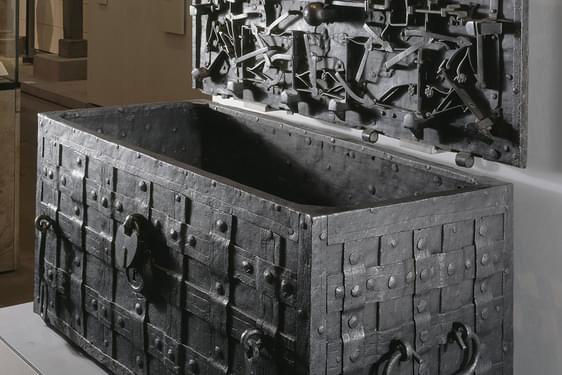 A metal chest decorated with a lattice weave. An open lid is hung on the wall next to the chest.  The inside of the lid has multiple metal objects with floral motifs attached to it.