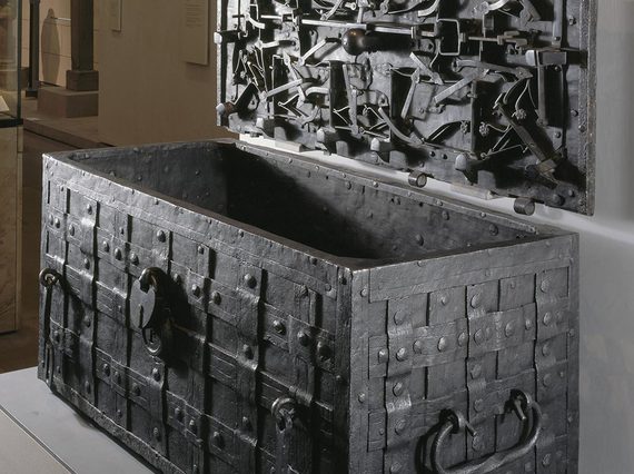 A metal chest decorated with a lattice weave. An open lid is hung on the wall next to the chest.  The inside of the lid has multiple metal objects with floral motifs attached to it.