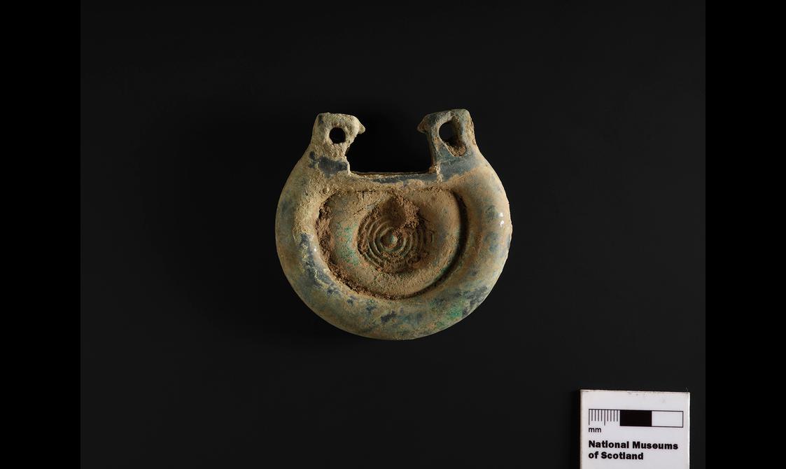 A bronze fitting with some soil still visible on its surface. It's rounded, with a concentric circle design.