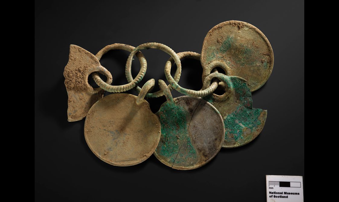 Three bronze rings linked together in a row. Five bronze disks are hanging from these rings, creating a rattle.