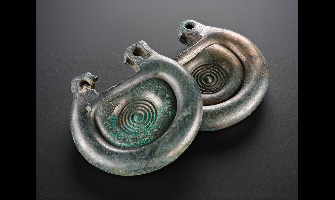 A bronze fitting that is silvery in colour with some green corrosion on the surface. It's rounded, with a concentric circle design.