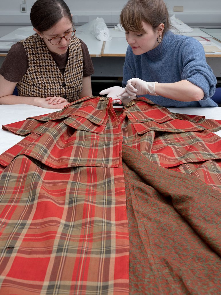 Two people seated at the end of the table examining a laid out tartan cape on a table.